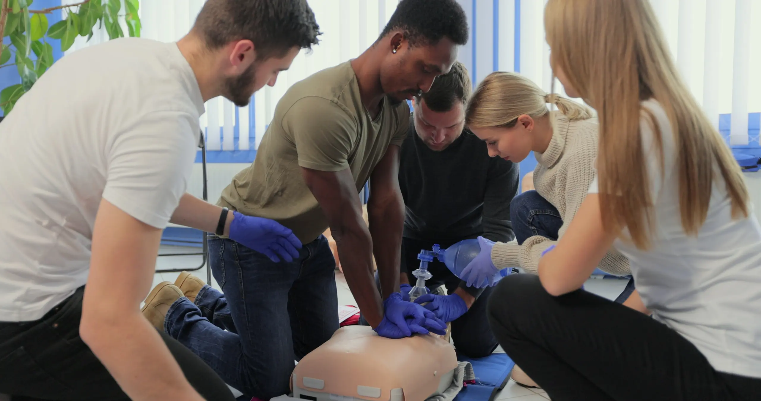 Five trainees wearing blue medical gloves practicing CPR techniques together on adult training mannequin during hands-on first aid certification class in bright training room