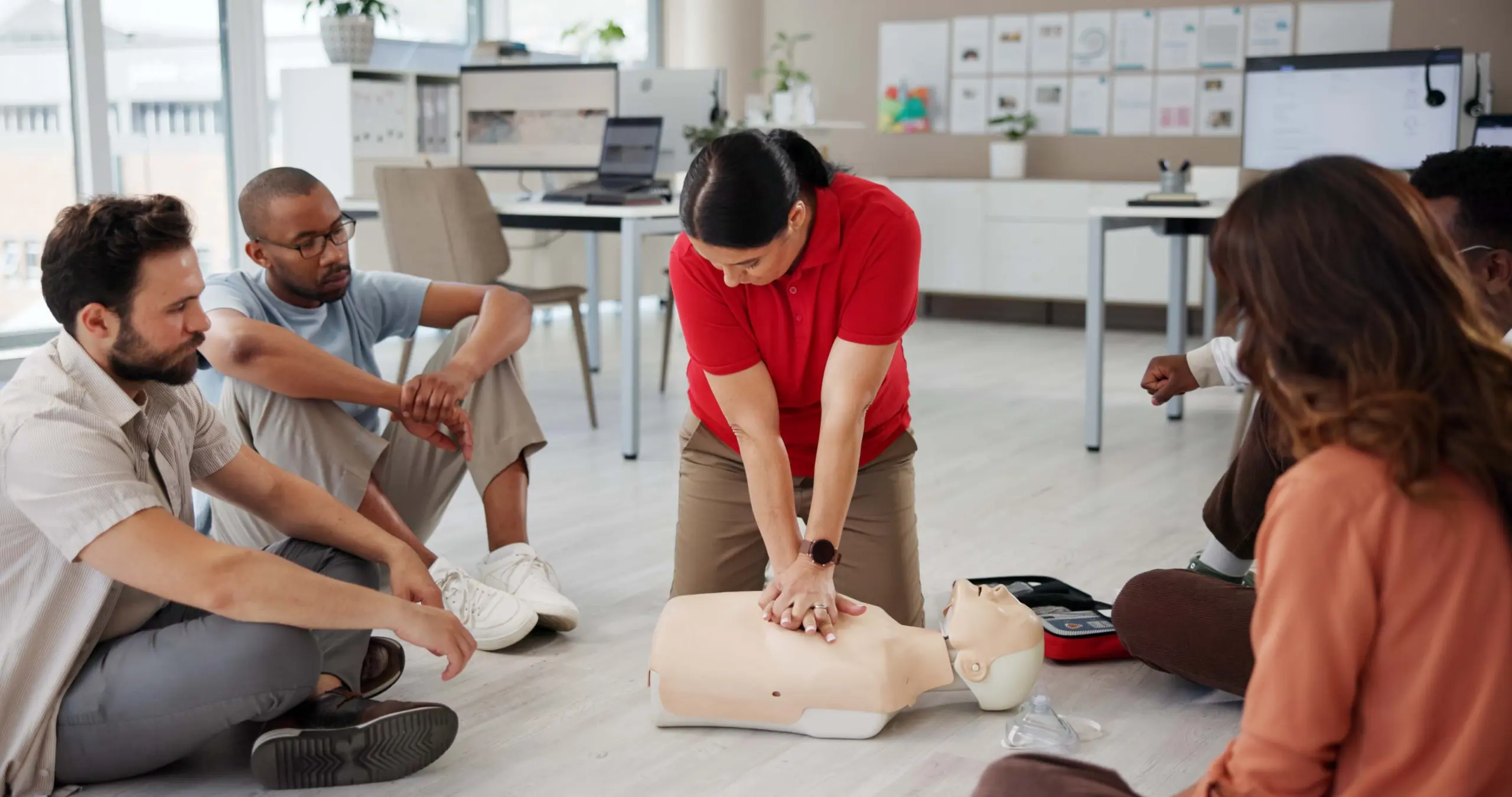 Instructor in red shirt demonstrating CPR chest compressions on training mannikin to group of four employees during workplace first aid training session in modern office