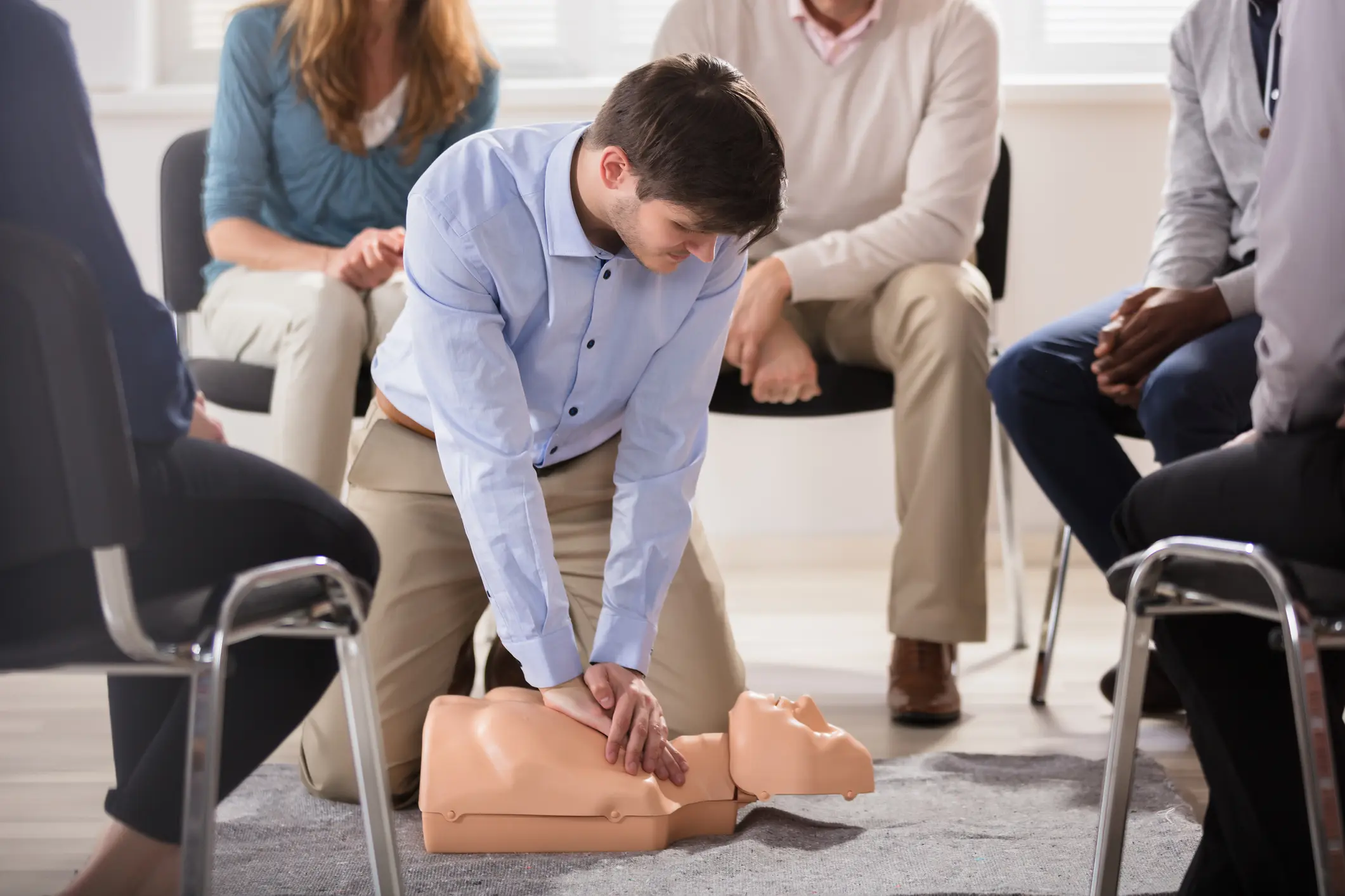 Man in blue shirt practicing CPR chest compressions on adult training mannequin while group of trainees observe from seated circle during first aid certification class in bright room