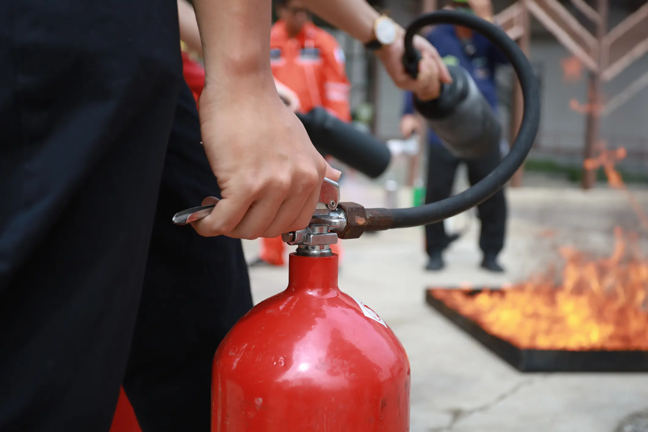 Close-up of trainee's hands operating red fire extinguisher during hands-on fire safety training with controlled flames and other participants practicing in background