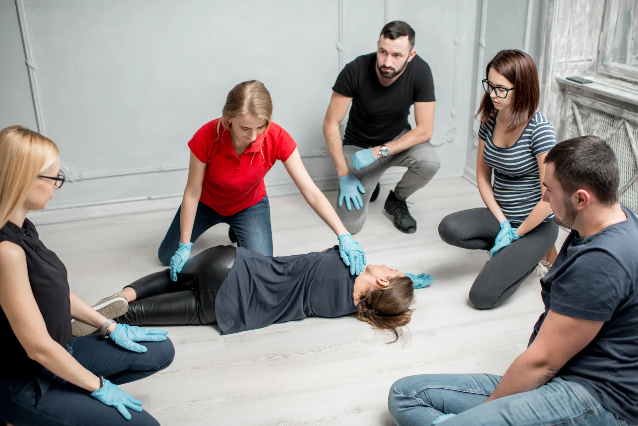 Instructor in red shirt demonstrating recovery position technique on mannequin while five trainees wearing blue medical gloves observe during first aid certification class