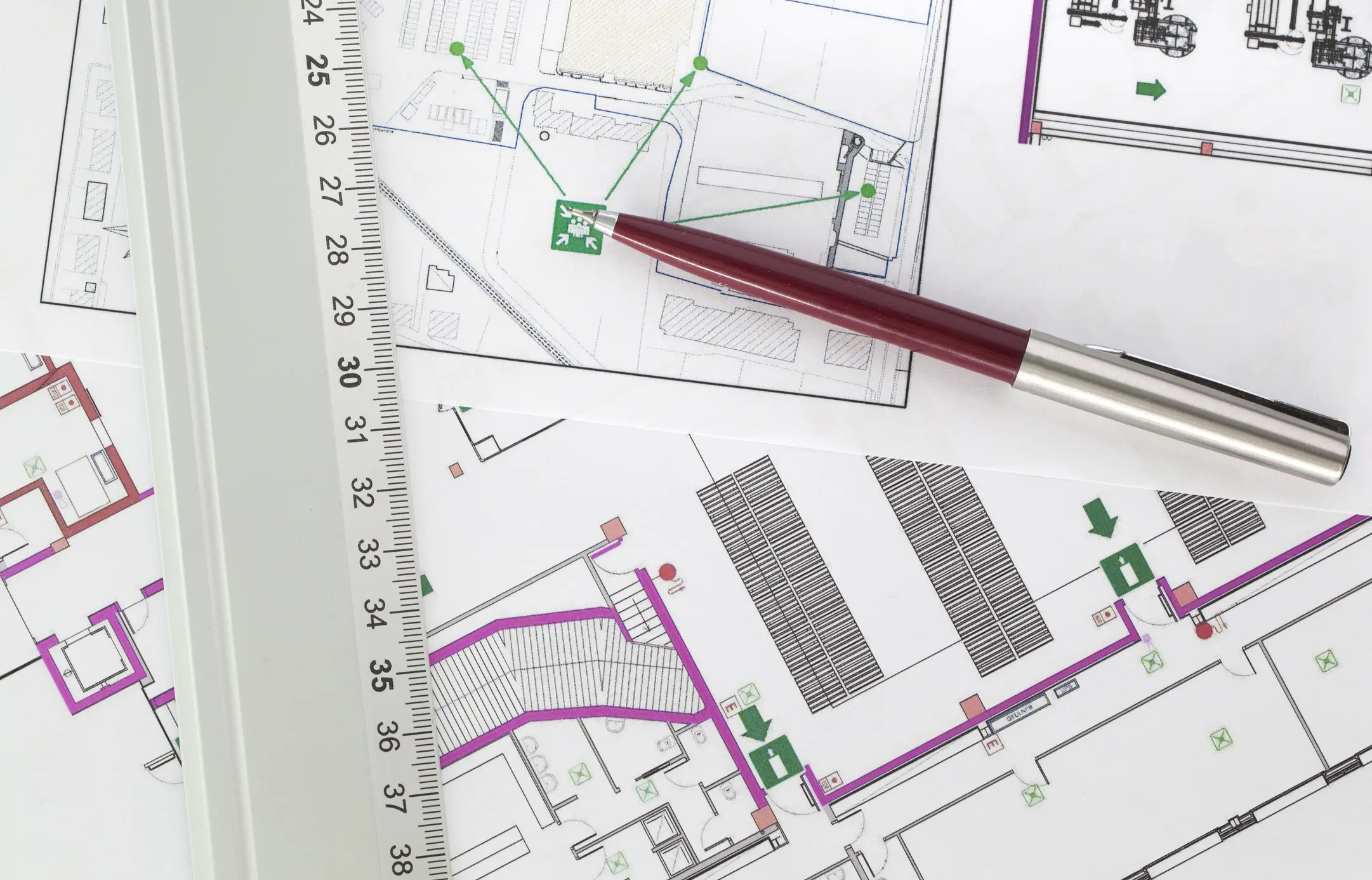 Fire safety evacuation floor plan showing emergency exit routes marked with green arrows and exit signs, with red pen and ruler on architectural blueprint